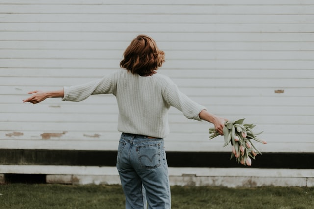 woman facing away from camera with arms spread wide, holding flowers to represent a confident woman who likes to be alone