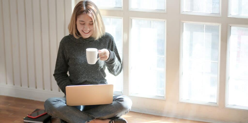 girl sitting on the floor by a window drinking coffee and using a laptop