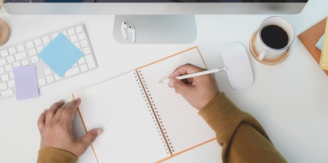 ariel view of desk with hands writing in notebook next to coffee and a keyboard