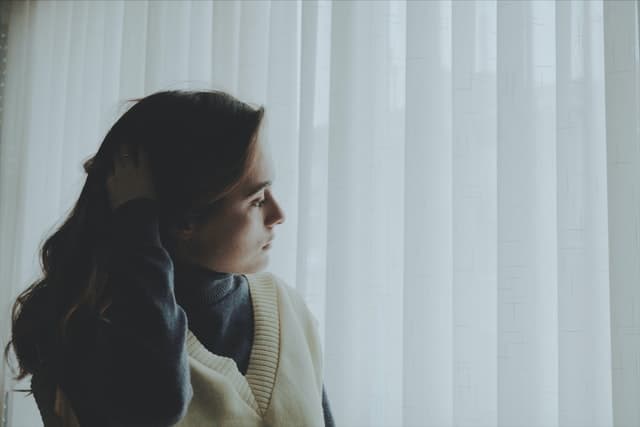 girl in sweater looking out window with curtains