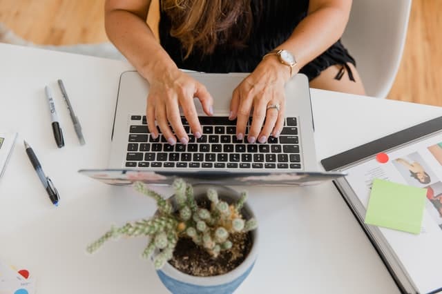 hands typing on a laptop at a desk with a plant and pens