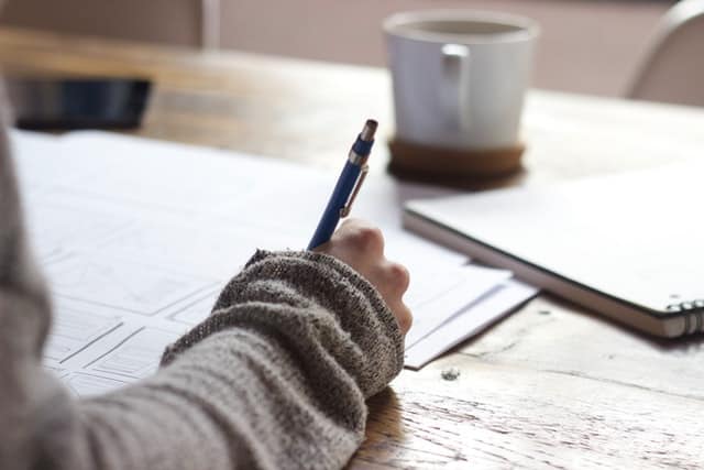 hand holding pen writing on paper next to notebook and coffee