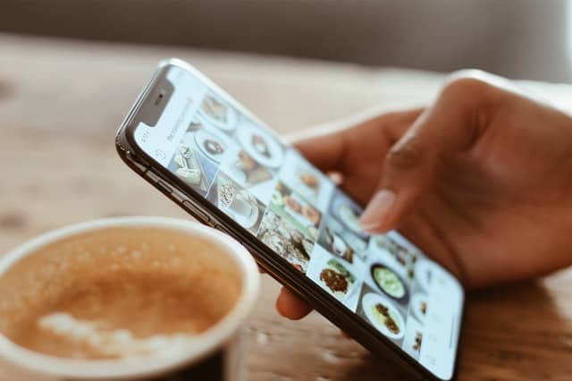 close up of hand using phone to scroll through pictures with coffee cup next to phone